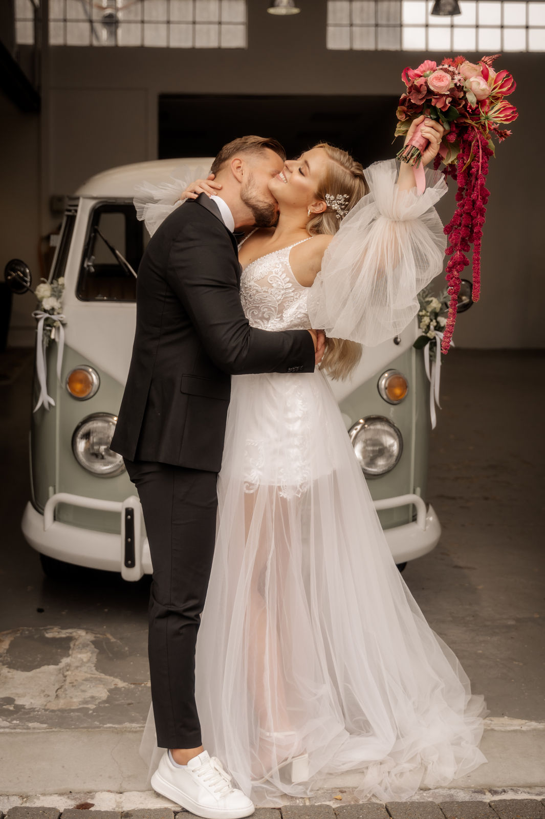 Glückliches Brautpaar küsst sich vor Vintage-Bus mit Brautstrauß